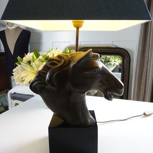 May include: A black lamp with a dark green lampshade and a horse head sculpture base. The horse head is made of a dark brown material and is facing to the right. The lamp is on a white surface.