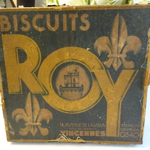 Op de afbeelding: Vintage doos van "Biscuits Roy" met een goud en zwart ontwerp. De doos heeft een kasteel illustratie en fleur-de-lis symbolen. De tekst op de doos luidt "Biscuits Roy", "14. Avenue de la Villa", "Face au Chateau", "Vincennes", "Téléphone Daumesnil 0540".