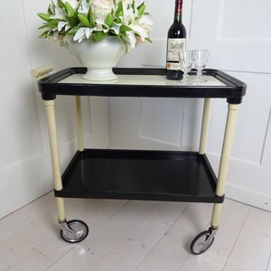 Può includere: Un carrello da servizio a due livelli nero e bianco con gambe e ruote color crema. Il carrello ha un piano superiore nero e uno scaffale inferiore nero. Una bottiglia di vino e due bicchieri sono sul piano superiore.