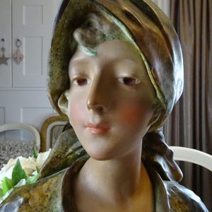 May include: A ceramic bust of a woman with a brown and green draped fabric head covering. The bust is painted with realistic skin tones and blush on the cheeks.