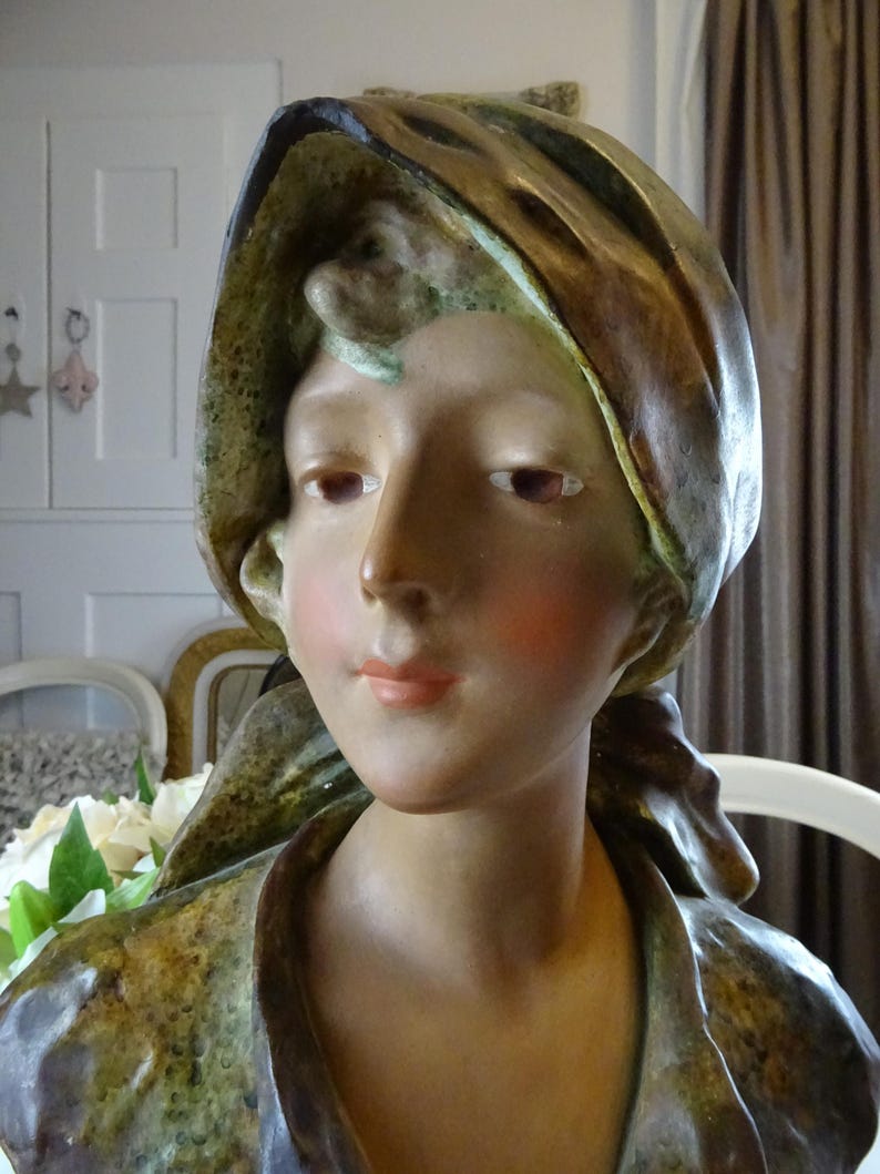 May include: A ceramic bust of a woman with a brown and green draped fabric head covering. The bust is painted with realistic skin tones and blush on the cheeks.