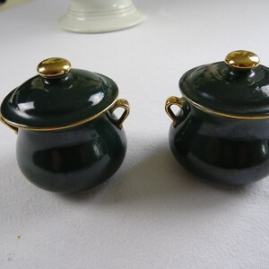 Può includere: Due piccoli vasi in ceramica verde con coperchi e manici dorati. I vasi hanno un bordo dorato intorno alla parte superiore.