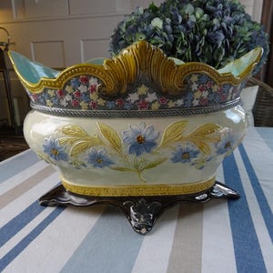 Op de afbeelding: Een keramieken schaal met een crèmekleurig lichaam, versierd met reliëf van blauwe bloemen en gele bladeren. De geschulpte rand heeft een bloemmotief in blauw, rood en geel, terwijl de donkerbruine voet op vier sierlijke pootjes rust.