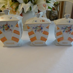 Könnte beinhalten: Set aus drei weißen Keramikbehältern mit orangefarbenen und goldenen Akzenten. Die Behälter haben Blumendesigns und sind mit "Farine", "Sucre" und "Cafe" beschriftet.