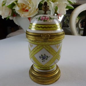 Op de afbeelding: Een porseleinen sieradendoosje met een geel en wit bloemenmotief. Het doosje heeft een scharnierend deksel en een gouden basis.