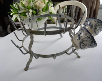 May include: A vintage metal cup drying rack with a distressed finish. The rack holds two decorative cups with blue floral patterns. The rack is supported by curved legs and has multiple arms for holding cups. The overall design is rustic and charming.