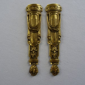 May include: A pair of ornate gold-colored metal decorative pieces with intricate designs. Each piece features a column-like structure with a draped fabric motif and a decorative finial at the bottom.