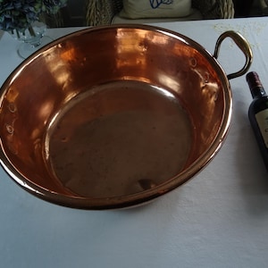 Op de afbeelding: Een grote, ronde kom van gepolijst koper met twee gebogen messing handgrepen die met klinknagels aan weerszijden zijn bevestigd. De kom heeft een glad, reflecterend binnenoppervlak en een brede opening. Een wijnfles staat naast de kom.
