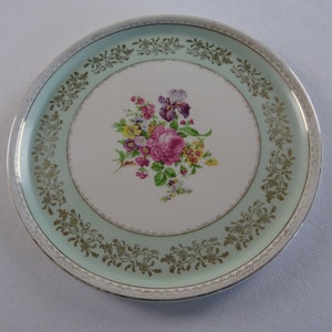May include: A white ceramic plate with a green and gold border. The centre of the plate features a floral design with pink, purple, and yellow flowers.