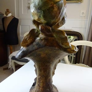 May include: A ceramic bust sculpture of a woman with a green and brown glaze. The woman is wearing a hat and her head is tilted down. The sculpture is on a pedestal.
