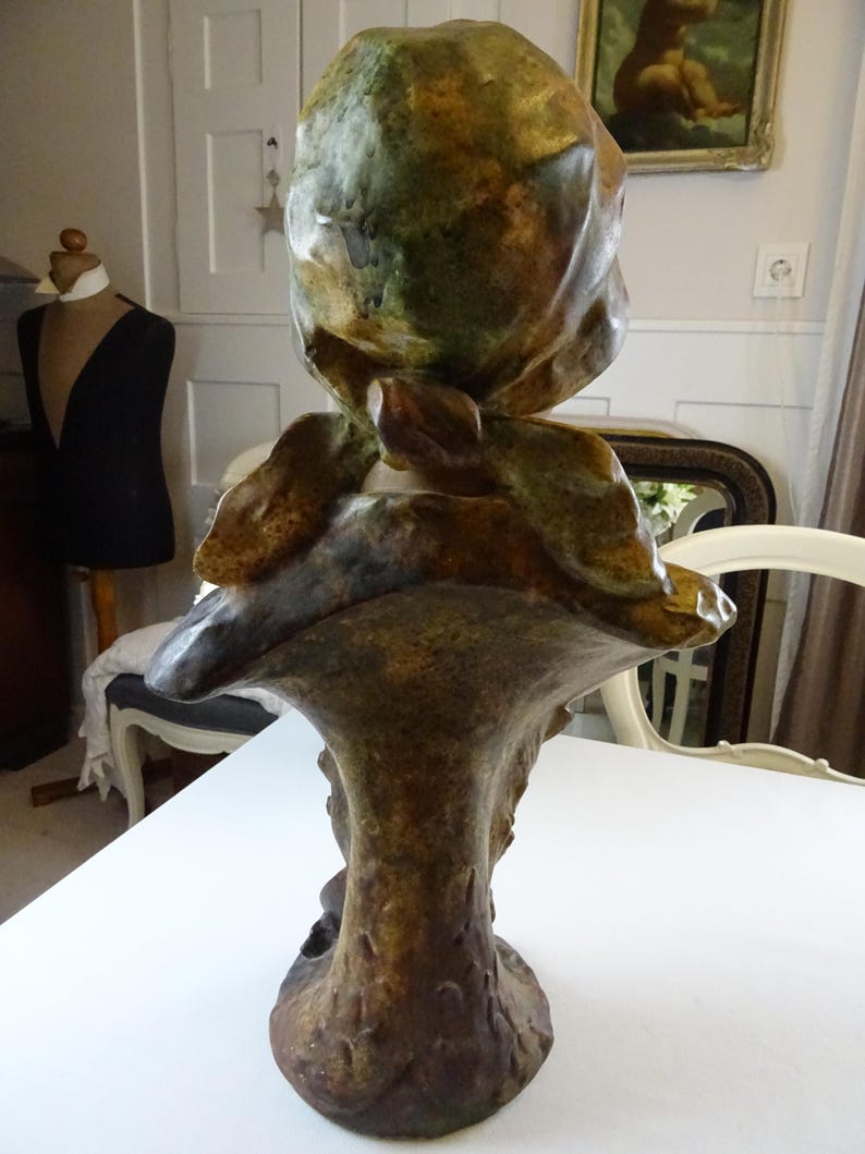 May include: A ceramic bust sculpture of a woman with a green and brown glaze. The woman is wearing a hat and her head is tilted down. The sculpture is on a pedestal.