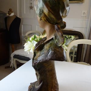 May include: A ceramic bust sculpture of a woman with a green and brown patterned head scarf. The bust is painted in shades of brown and green, and the woman's face is turned to the side.