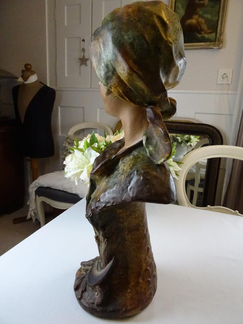 May include: A ceramic bust sculpture of a woman with a green and brown patterned head scarf. The bust is painted in shades of brown and green, and the woman's face is turned to the side.