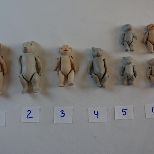 Könnte beinhalten: Eine Sammlung antiker Bisquitpuppen, jede mit beweglichen Gliedmaßen und bemalten Gesichtszügen. Die Puppen sind in verschiedenen Posen und Größen, mit unterschiedlichen Hauttönen und Gesichtsausdrücken. Nummerierte Karten sind unter jeder Puppe platziert.