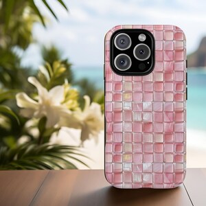 May include: A pink and white mosaic tile phone case with a black border. The case features a camera cutout. The background is a blurred image of a beach with white flowers and green foliage.