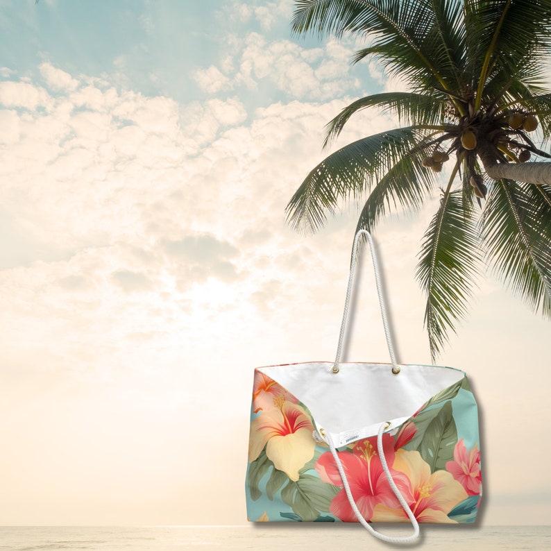 May include: A white tote bag with a tropical floral print featuring pink, red, and yellow hibiscus flowers on a light blue background. The bag has white rope handles.