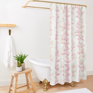 Op de afbeelding: Een wit douchegordijn met een roze bloemenpatroon. Het gordijn heeft een gouden douchestang en hangt in een badkamer met een wit bad en een houten kruk.