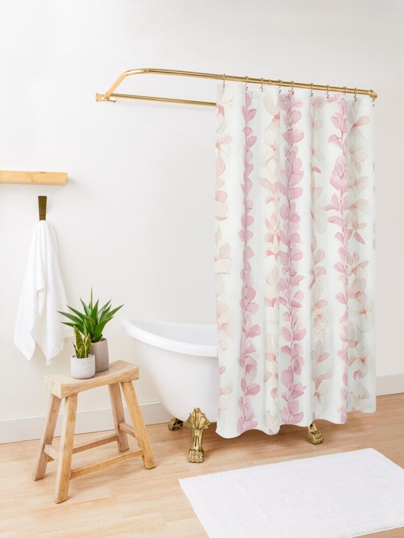Op de afbeelding: Een wit douchegordijn met een roze bloemenpatroon. Het gordijn heeft een gouden douchestang en hangt in een badkamer met een wit bad en een houten kruk.