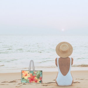 May include: A woman wearing a white swimsuit and a straw hat sits on a sandy beach, looking out at the ocean. A floral tote bag with a white handle sits on the sand in front of her.