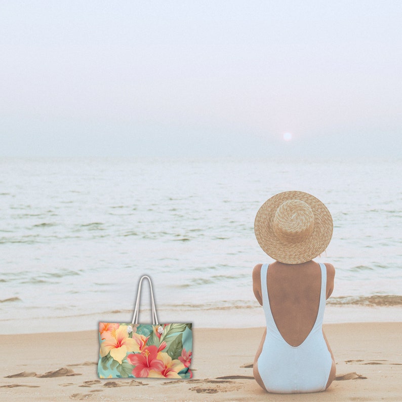 May include: A woman wearing a white swimsuit and a straw hat sits on a sandy beach, looking out at the ocean. A floral tote bag with a white handle sits on the sand in front of her.