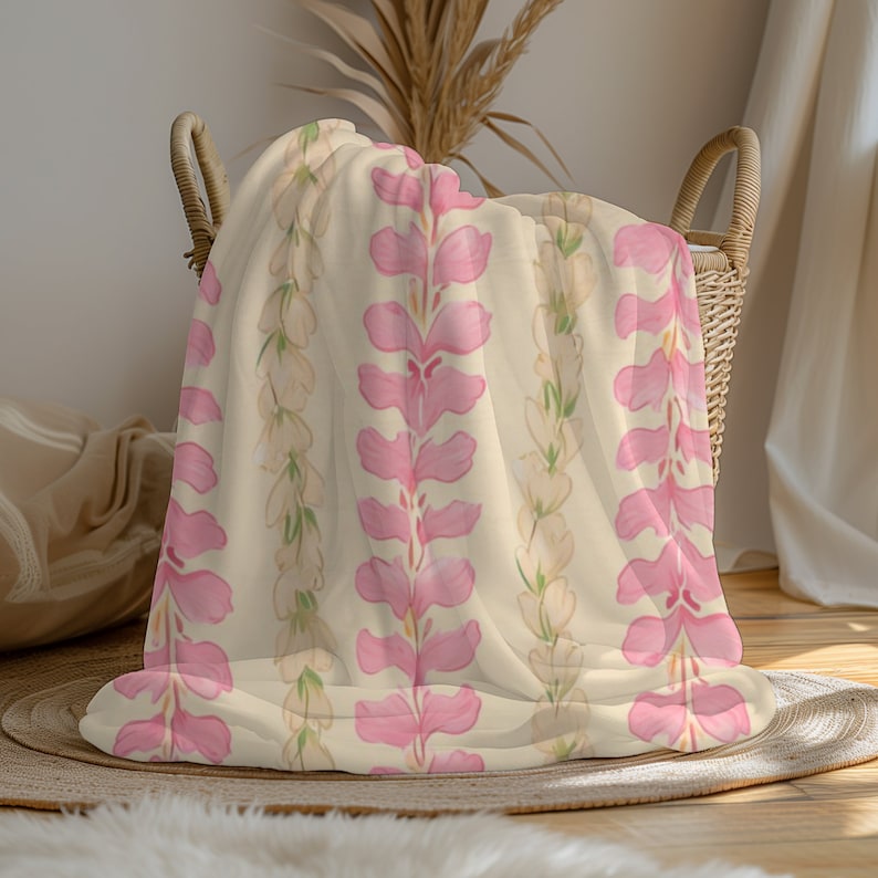 May include: A soft, cream-colored throw blanket with a repeating pattern of pink and white floral designs. The blanket is folded and displayed in a wicker basket.