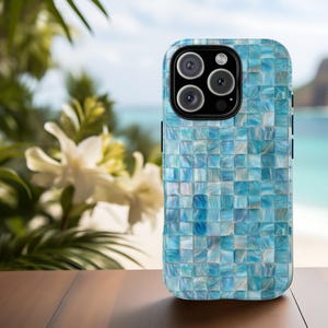 May include: A smartphone case with a mosaic tile design in shades of blue and white. The case has a glossy finish and a black camera module. The background shows a beach scene with white flowers and a wooden table.
