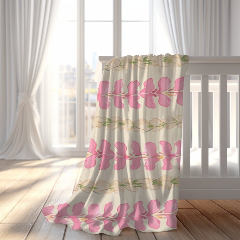 May include: A pink and white floral baby blanket with a repeating pattern of pink flowers and white leaves. The blanket is draped over a white crib.