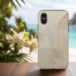 May include: A beige phone case featuring a textured surface and an abstract design of curved lines and arches. The case is on a wooden surface with a blurred tropical background of flowers, ocean, and sky.