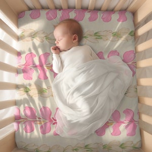 May include: A baby sleeping in a crib with a pink floral patterned sheet. The baby is wearing a white onesie and is swaddled in a white blanket.