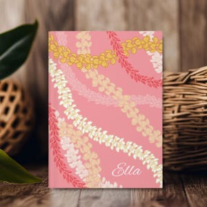 Può includere: Un taccuino rosa con un motivo floreale con ghirlande gialle, bianche e rosa. La parola "Ella" è scritta in corsivo nell'angolo in basso a destra. Il taccuino è rettangolare e sembra essere su una superficie di legno.