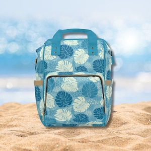 Puede incluir: Una mochila azul y blanca con estampado de hojas tropicales, con un bolsillo frontal grande y correas ajustables.