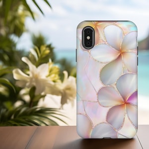 Puede incluir: Una funda de teléfono con un diseño floral con detalles en blanco, rosa y dorado. La funda tiene un acabado brillante y un patrón de flores de plumeria estilizadas. El fondo muestra una escena de playa borrosa con flores blancas.