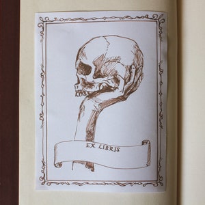 Puede incluir: Un dibujo a tinta marr&oacute;n de una mano sosteniendo una calavera humana con un borde decorativo y el texto "Ex Libris".