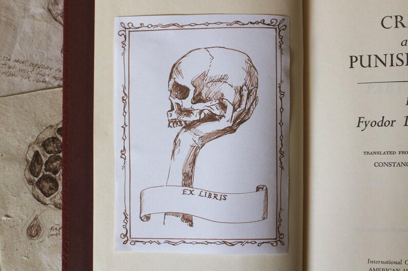 Puede incluir: Un dibujo a tinta marr&oacute;n de una mano sosteniendo una calavera humana con un borde decorativo y el texto "Ex Libris".