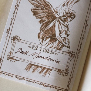 Puede incluir: Un dibujo a tinta marr&oacute;n de un &aacute;ngel con las alas extendidas, sosteniendo un libro. El dibujo est&aacute; enmarcado con un borde decorativo e incluye el texto "Ex Libris" y "Paul Macadamia".