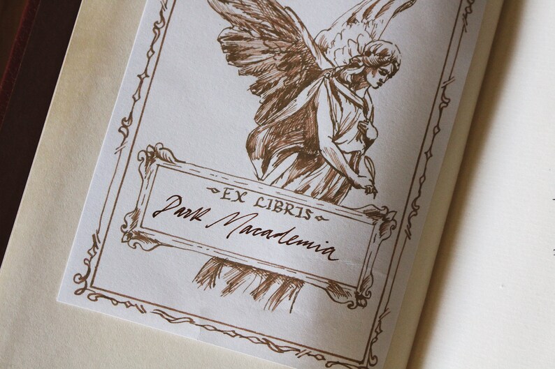 Puede incluir: Un dibujo a tinta marr&oacute;n de un &aacute;ngel con las alas extendidas, sosteniendo un libro. El dibujo est&aacute; enmarcado con un borde decorativo e incluye el texto "Ex Libris" y "Paul Macadamia".