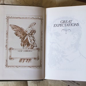 Puede incluir: Un libro vintage titulado "Grandes esperanzas" de Charles Dickens. La portada presenta un dibujo de un &aacute;ngel con alas y un borde decorativo. El libro est&aacute; abierto en la p&aacute;gina del t&iacute;tulo, que tiene una imagen descolorida de la cara de un hombre.
