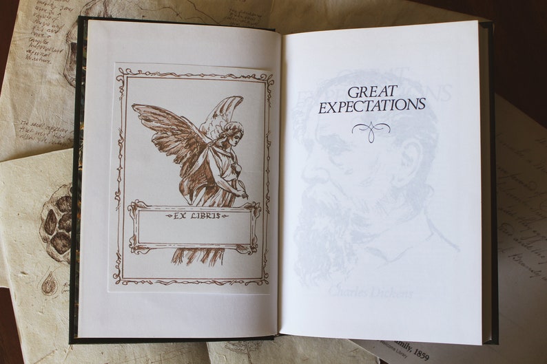 Puede incluir: Un libro vintage titulado "Grandes esperanzas" de Charles Dickens. La portada presenta un dibujo de un &aacute;ngel con alas y un borde decorativo. El libro est&aacute; abierto en la p&aacute;gina del t&iacute;tulo, que tiene una imagen descolorida de la cara de un hombre.