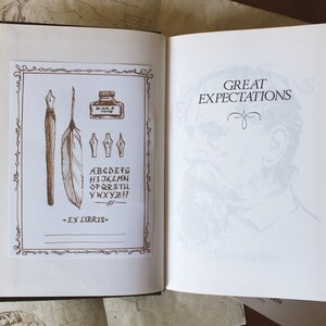 Puede incluir: Un libro abierto con fondo blanco. La p&aacute;gina izquierda tiene un borde decorativo con una pluma, una pluma, un tintero y un juego de plumillas. La p&aacute;gina derecha muestra el t&iacute;tulo "Great Expectations" en texto negro. El libro est&aacute; sobre una superficie de madera marr&oacute;n.
