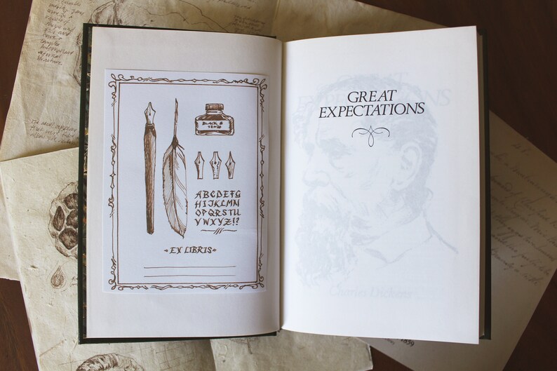 Puede incluir: Un libro abierto con fondo blanco. La p&aacute;gina izquierda tiene un borde decorativo con una pluma, una pluma, un tintero y un juego de plumillas. La p&aacute;gina derecha muestra el t&iacute;tulo "Great Expectations" en texto negro. El libro est&aacute; sobre una superficie de madera marr&oacute;n.