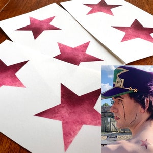 May include: White cards featuring a large, magenta star cutout design. The cards are arranged on a wooden surface. A person is wearing a purple hat with a green brim and a gold emblem, and has a star tattoo on their shoulder.