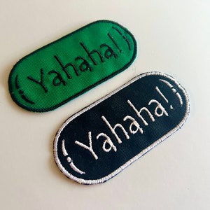 Peut inclure: Deux écussons brodés de forme ovale. L'un est vert avec des coutures noires et le mot "Yahaha!" en blanc. L'autre est bleu marine avec des coutures blanches et le même texte. Les deux ont une bordure blanche.