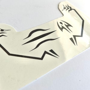 Puede incluir: Una pegatina de color crema con diseños negros de estilo tribal. La pegatina presenta un ojo, líneas curvas y formas geométricas. La pegatina está sobre una superficie blanca y tiene un acabado brillante.