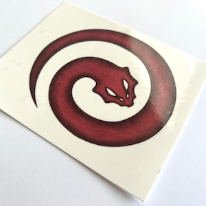 May include: A sticker featuring a stylized, burgundy-colored serpent in a circular design. The serpent's head is visible, with two white eye shapes. The sticker is on a white background.