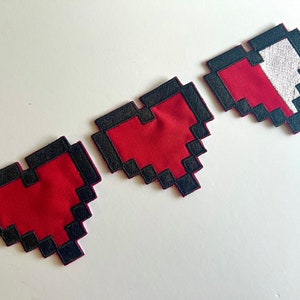 Op de afbeelding: Drie gepixelde hartvormige patches met een rode kern en zwarte rand. Het hart rechts heeft een wit gedeelte, wat een gezondheidsbalk suggereert. Deze patches zijn waarschijnlijk voor kleding of accessoires.