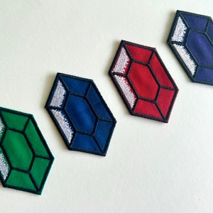 May include: Four embroidered patches in the shape of faceted gems. The patches are green, red, and two shades of blue. Each gem has a black outline and white stitching. These patches are suitable for clothing or accessories.