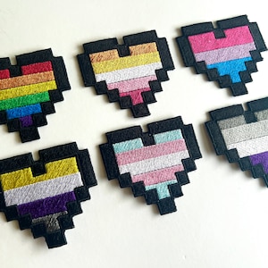 Puede incluir: Seis parches en forma de corazón pixelados con bordes negros. Cada corazón presenta un diseño de bandera del orgullo LGBTQ+ diferente, incluyendo banderas arcoíris, bisexuales, transgénero, no binarias, pansexuales y asexuales. Los parches están bordados.