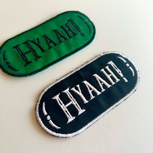 Peut inclure: Deux écussons brodés de forme ovale. L'un est vert avec le texte noir "HYAAH!". L'autre est bleu marine avec le texte blanc "HYAAH!". Les deux écussons ont une bordure cousue.