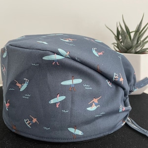 May include: A dark grey scrub cap featuring a repeating pattern of surfers on light blue surfboards. The surfers are shown in different positions, some standing and others riding waves. The cap has a tie closure.