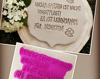 May include: A pink rubber stamp with the text "Unser Garten ist nicht ungepflegt! Er ist Lebensraum fur Insekten!"  The stamp is on a wooden surface with a white ceramic tile with the same text.
